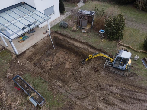 Vue a&eacute;rienne d'un chantier de terrassement r&eacute;sidentiel avec pelleteuse jaune creusant une excavation en forme de L pr&egrave;s d'une piscine couverte