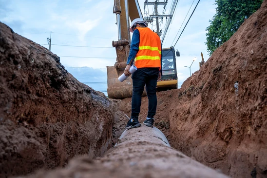 Chantier de travaux publics en extérieur avec ouvrier en gilet haute visibilité orange inspectant une canalisation dans une tranchée