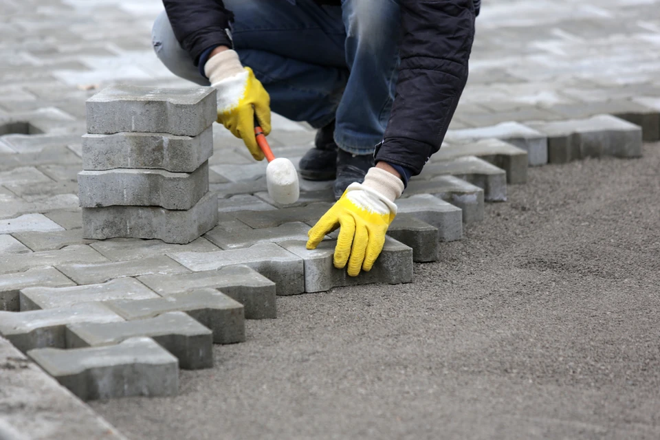 Ouvrier en tenue de travail posant des pavés en béton sur lit de sable, travaillant à genoux avec gants de protection jaunes et marteau à embout plastique blanc